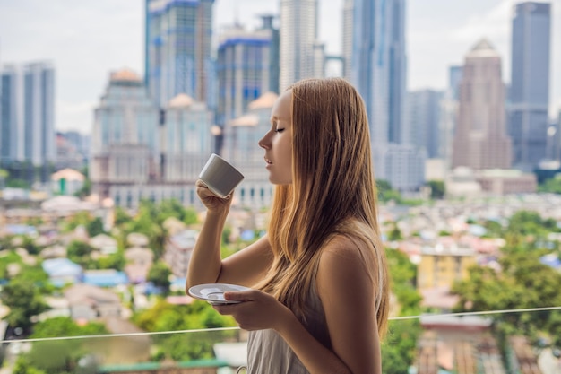 Młoda kobieta pije kawę rano na balkonie z widokiem na duże miasto i drapacze chmur