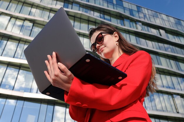 Młoda kobieta pisze na laptopie przed budynkiem biurowymPracownik biurowy pracuje na zewnątrz