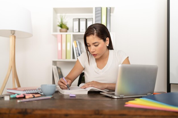 Młoda kobieta pisze w zeszycie z laptopem i filiżanką kawy na boku