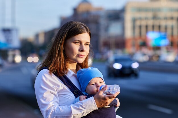 Młoda kobieta podaje dziecku napój w chuście na ulicy