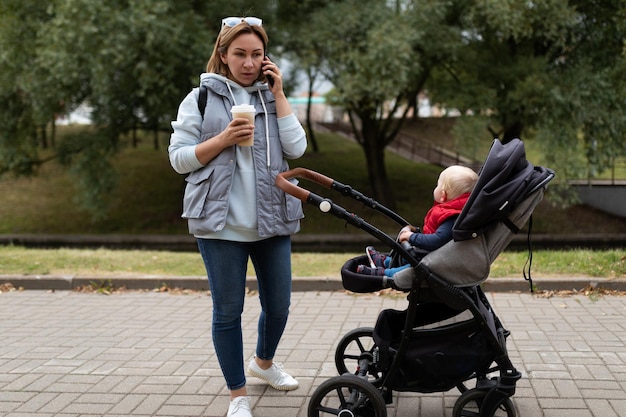 Młoda kobieta podczas spaceru z dzieckiem i wózkiem rozmawia przez telefon komórkowy i pije kawę w a