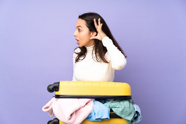Młoda kobieta podróżnik z walizką pełną ubrań na odosobnionej fioletowej ścianie słuchając czegoś, kładąc rękę na uchu