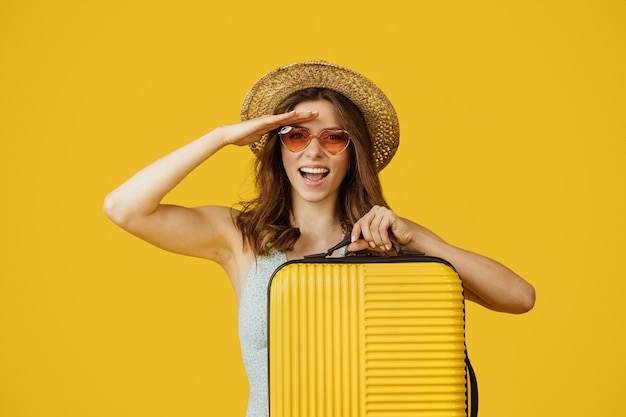 Młoda kobieta podróżująca w słomkowym kapeluszu niosąca walizkę i patrząca na kamerę stojącą na żółtym tle studia