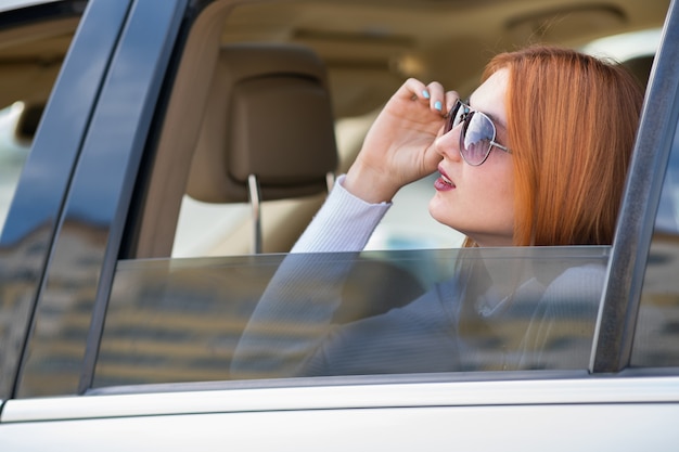 Młoda Kobieta Podróżuje Samochodem Z Czerwonymi Włosy I Słońca Szkłami. Pasażer Patrząc Przez Tylne Okno Taksówki W Mieście.
