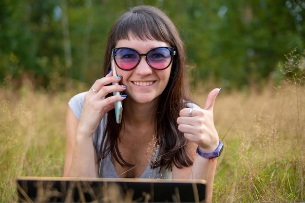 Młoda kobieta pokazuje kciuk w górę, dzwoniąc przez telefon i uśmiechając się szczęśliwie na letnim trawniku na zewnątrz
