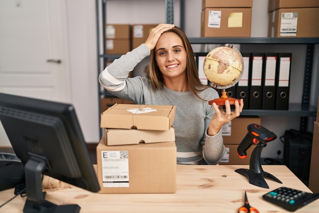 Młoda kobieta pracująca w małym biznesie e-commerce trzymająca światową piłkę zestresowana i sfrustrowana ręką na głowie, zdziwioną i wściekłą twarzą
