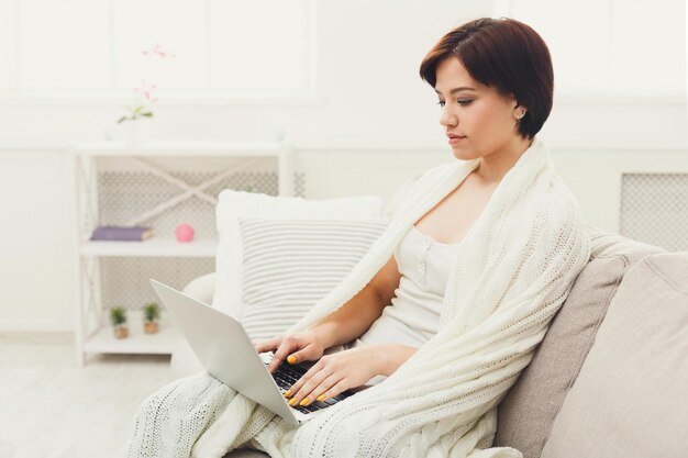 Młoda kobieta pracuje na laptopie. Rozważna kobieta przeglądająca nowe wiadomości w sieciach społecznościowych, siedząca na kanapie, owinięta białym kocem, kopiująca przestrzeń. Koncepcja przytulności i technologii.