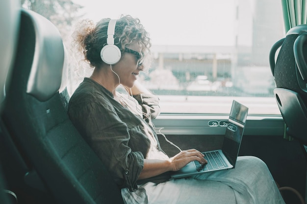 Młoda kobieta pracuje na laptopie z zestawem słuchawkowym siedzącym na siedzeniu autobusu Podróżujący biznesowy styl życia kobiety Nowoczesna dama uśmiecha się i cieszy się podróżą jako pasażer w transporcie publicznym pojazdu Technologia online