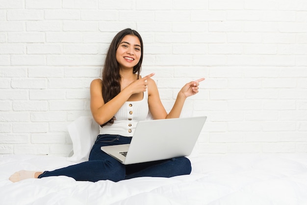 Młoda kobieta pracuje z laptopem na łóżku podekscytowany, wskazując palcami wskazującymi daleko