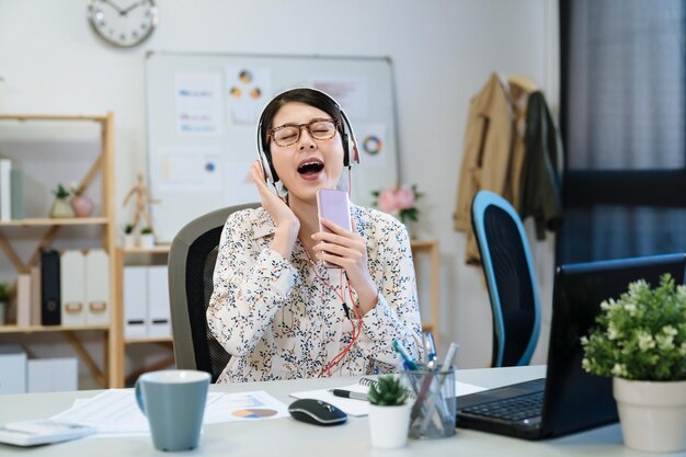 Młoda kobieta projektantka mody w słuchawkach siedzi przy biurku zdumiewająco za pomocą telefonu komórkowego śpiewa w pracy w nowoczesnym biurze. szczęśliwa krawiecka w słuchawkach i trzymająca telefon komórkowy jako mikrofon w studio