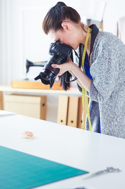 Młoda kobieta projektantka stojąca w pobliżu miejsca pracy i fotografująca je aparatem cyfrowym