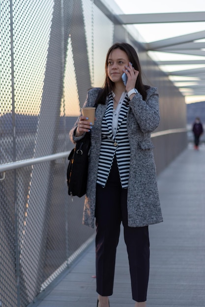 Młoda kobieta przechodząca przez most dla pieszych, rozmawiająca przez telefon i trzymająca filiżankę kawy