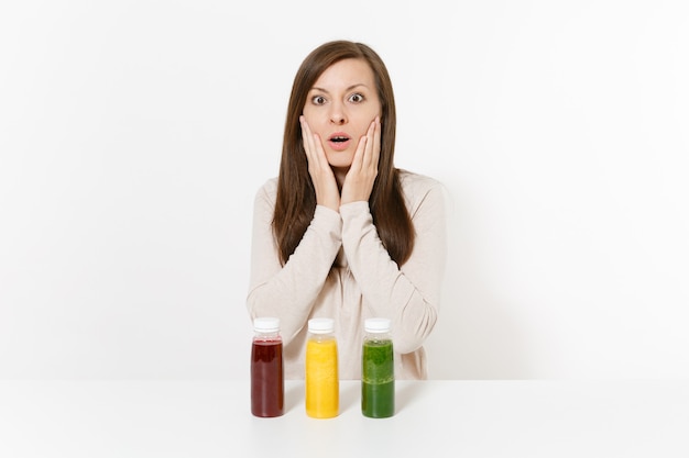 Zdjęcie młoda kobieta przy stole z zielonymi, czerwonymi i żółtymi koktajlami detox w butelkach na białym tle. prawidłowe odżywianie, napój wegetariański, zdrowy styl życia, koncepcja diety. obszar z miejscem na kopię.