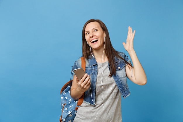 Młoda kobieta radosna studentka w dżinsowe ubrania z plecakiem, słuchawki rozłożone ręce słuchania muzyki trzymać telefon komórkowy na białym tle na niebieskim tle. Edukacja na studiach. Skopiuj miejsce na reklamę.