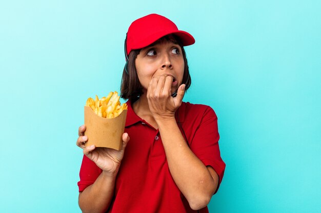 Młoda kobieta rasy mieszanej fast food restauracja pracownik gospodarstwa frytki na białym tle na niebieskim tle gryząc paznokcie, nerwowy i bardzo niespokojny.