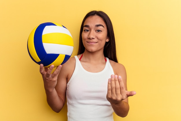 Młoda kobieta rasy mieszanej gra w siatkówkę na plaży na białym tle na żółtym tle wskazując palcem na ciebie, jakby zapraszając się bliżej.