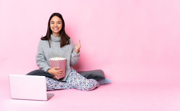 Młoda kobieta rasy mieszanej jedzenie popcornu podczas oglądania filmu na laptopie, wskazując na świetny pomysł