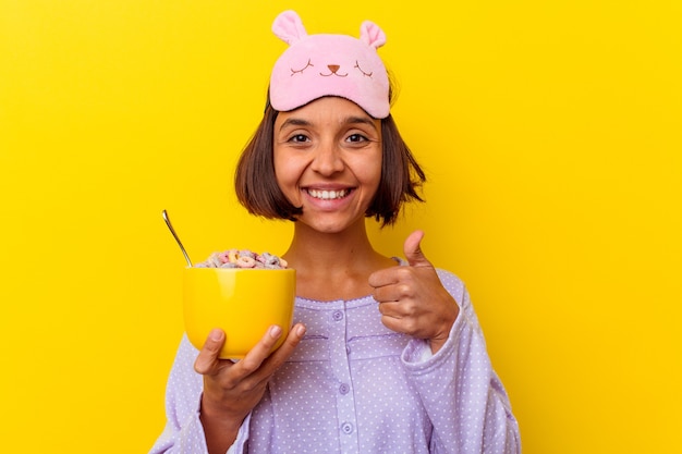 Młoda Kobieta Rasy Mieszanej Jedzenie Zbóż Ubrana W Pijama Na Białym Tle Na żółtym Tle Uśmiechając Się I Podnosząc Kciuk Do Góry