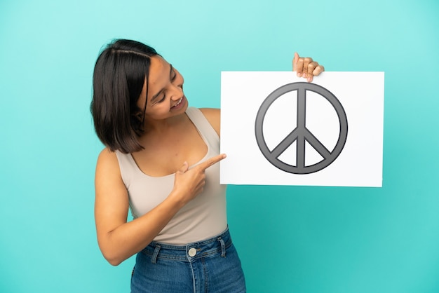 Młoda kobieta rasy mieszanej na białym tle na niebieskim tle trzymająca tabliczkę z symbolem pokoju i wskazującą go