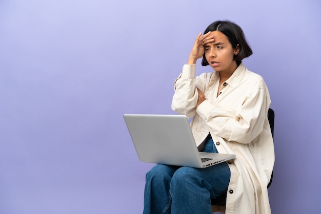 Młoda kobieta rasy mieszanej siedzi na krześle z laptopem na białym tle na fioletowym tle, patrząc daleko ręką, aby coś wyglądać