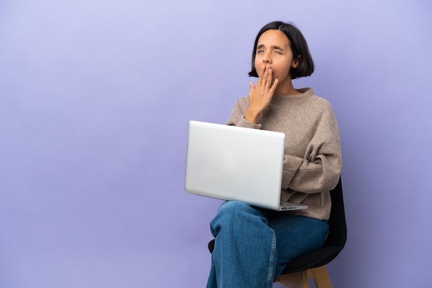 Młoda kobieta rasy mieszanej siedzi na krześle z laptopem na białym tle na fioletowym tle ziewanie i obejmujące szeroko otwarte usta ręką