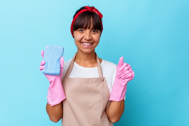 Młoda kobieta rasy mieszanej sprzątająca dom na białym tle na niebieskim tle, uśmiechnięta i podnosząca kciuk w górę