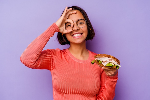 Młoda kobieta rasy mieszanej trzymając kanapkę na białym tle na fioletowej ścianie podekscytowany utrzymując ok gest na oko.