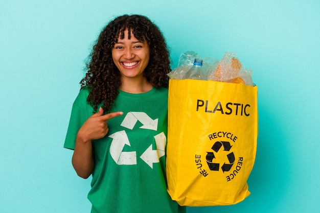 Młoda kobieta rasy mieszanej, trzymając plastikową torbę z recyklingu odizolowaną na niebieskiej ścianie, uśmiechając się i wskazując na bok, pokazując coś w pustym miejscu.