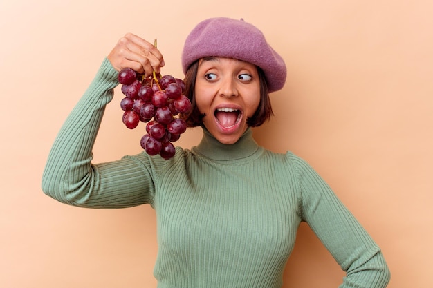 Młoda Kobieta Rasy Mieszanej Trzymając Winogrona Na Białym Tle Na Beżowej ścianie