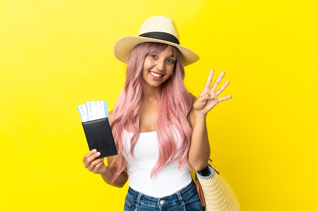Młoda kobieta rasy mieszanej trzymająca paszport i torbę plażową na białym tle na żółtym tle szczęśliwa i licząca cztery palcami