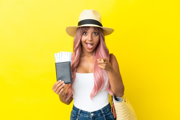 Młoda kobieta rasy mieszanej trzymająca paszport i torbę plażową na żółtym tle zaskoczona i wskazująca przód