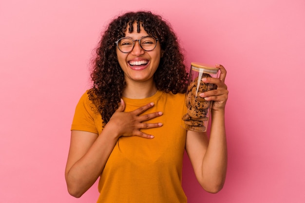 Młoda kobieta rasy mieszanej trzymająca słoik z ciasteczkami na różowej ścianie śmieje się głośno trzymając rękę na piersi.