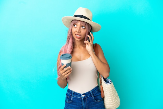 Młoda kobieta rasy mieszanej trzymająca torbę plażową z pamelą odizolowaną na niebieskim tle, trzymającą kawę na wynos i telefon komórkowy