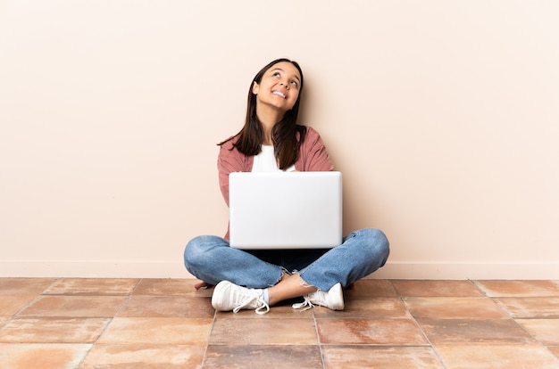 Młoda kobieta rasy mieszanej z laptopem siedząc na podłodze patrząc w górę uśmiechając się