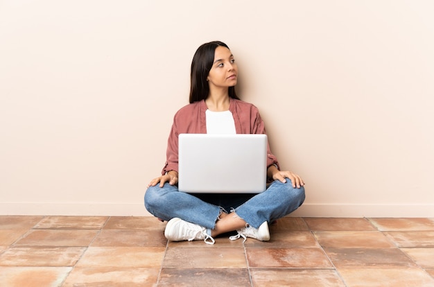 Młoda kobieta rasy mieszanej z laptopem siedzi na podłodze, trzymając ręce skrzyżowane
