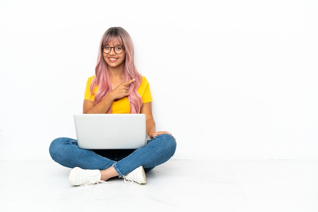 Młoda kobieta rasy mieszanej z laptopem z różowymi włosami siedzi na podłodze na białym tle, wskazując na bok, aby zaprezentować produkt