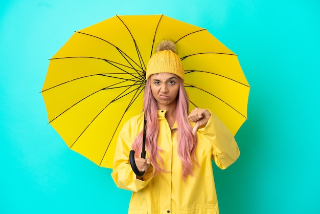 Zdjęcie młoda kobieta rasy mieszanej z płaszczem przeciwdeszczowym i parasolem pokazującym kciuk w dół z negatywnym wyrazem twarzy