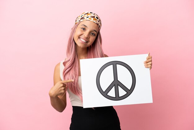 Zdjęcie młoda kobieta rasy mieszanej z różowymi włosami na różowym tle trzymająca tabliczkę z symbolem pokoju i wskazującą go