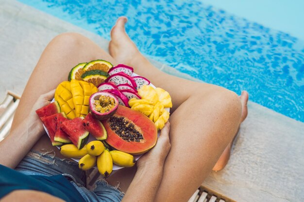 Młoda kobieta relaks i jedzenie talerz owoców przy hotelowym basenie. Egzotyczna letnia dieta. Zdjęcie nóg ze zdrową żywnością przy basenie, widok z góry z góry. Tropikalny styl życia na plaży