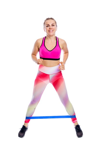 Młoda kobieta robi ćwiczenia nóg z gumką, stojąc Uśmiechnięta piękna kobieta w jasnej odzieży sportowej Aktywność sportowa i zdrowie Pojedynczo na białym tle Pełna wysokość Widok z przodu