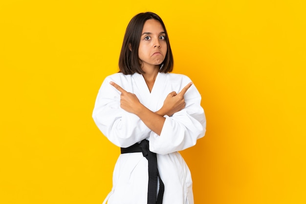 Młoda kobieta robi karate na białym tle na żółtej ścianie, wskazując na boczne, mając wątpliwości