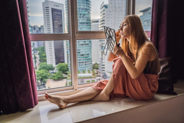 Młoda kobieta robi makijaż siedząc przy oknie z panoramicznym widokiem na wieżowce i duże miasto