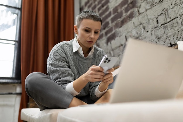 Młoda kobieta robi zakupy online za pomocą laptopa i telefonu komórkowego, siedząc na kanapie w domu