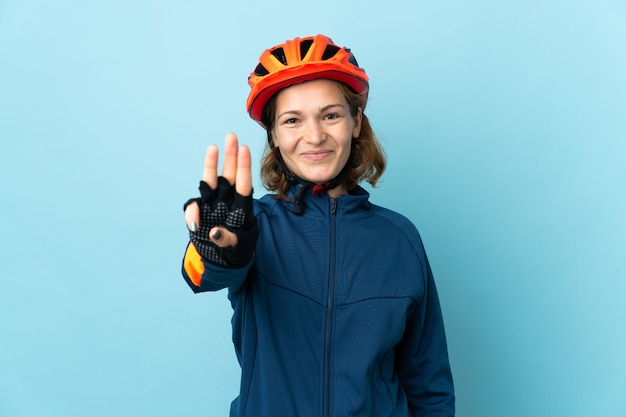 Młoda kobieta rowerzysta na białym tle na niebieskim tle szczęśliwa i licząc trzy palcami