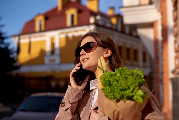 Młoda kobieta rozmawia przez telefon i trzyma paczkę z zakupami w rękach na tle domu