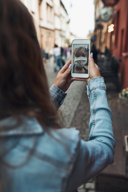 Młoda kobieta rozmawiająca przez połączenie wideo podczas spaceru po centrum miasta w masce twarzy, aby uniknąć wirusa
