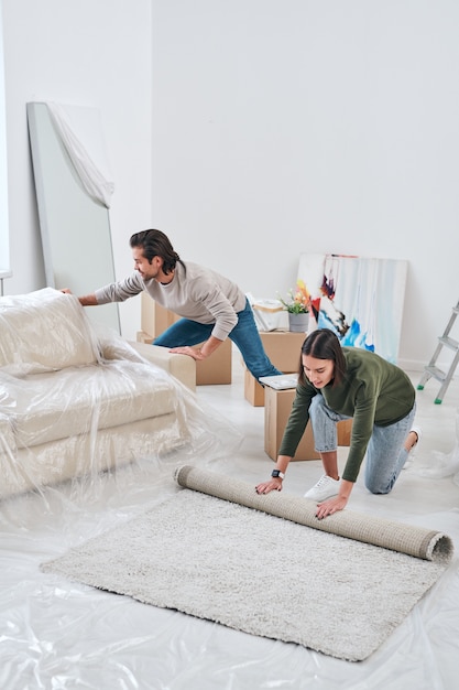 Młoda kobieta rozwija dywan na podłodze w salonie, podczas gdy jej mąż usuwa nową kanapę w pobliżu