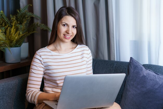 Młoda kobieta siedząca na kanapie, pracująca przy laptopie, przeglądająca internet lub czytająca wiadomości z domu happy fem