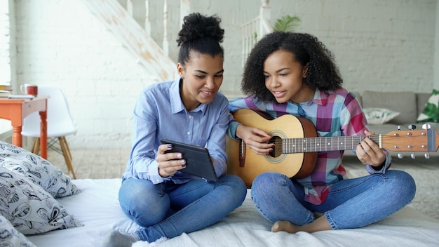 młoda kobieta siedząca na łóżku z tabletem i ucząca swoją nastoletnią siostrę gry na gitarze akustycznej