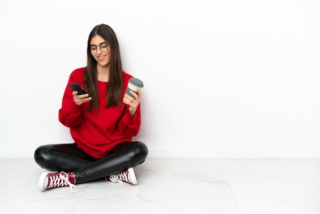 Młoda kobieta siedząca na podłodze na białym tle trzymająca kawę na wynos i telefon komórkowy